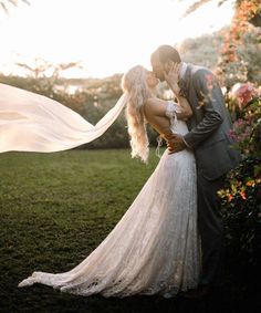 a bride and groom kissing in front of a flowered bush with their veil blowing in the wind