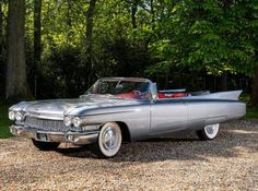 an old silver car parked in front of some trees and gravel with its hood up