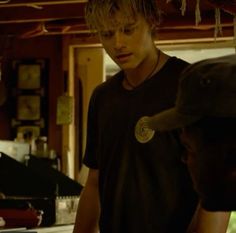 two men standing in a kitchen looking at something on the counter and one is wearing a hat