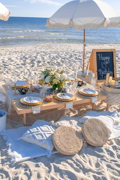 a table set up on the beach for a birthday party with an umbrella over it