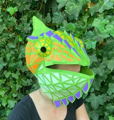 a woman wearing a paper fish mask in front of some bushes and ivys with her eyes closed