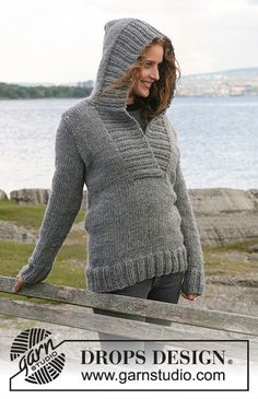 a woman wearing a hooded sweater standing on a wooden fence by the water with her hands in her pockets