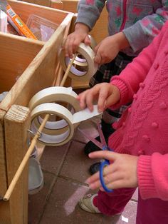 two children are playing with craft supplies in a wooden box on the floor, while another child is holding scissors and tape