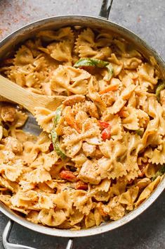 a skillet filled with pasta and vegetables on top of a gray counter next to a wooden spoon