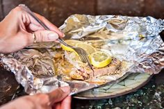 two hands are holding a fork and knife over some foil wrapped food on a plate