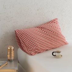 a red and white checkered pillow sitting on top of a bed next to a cup