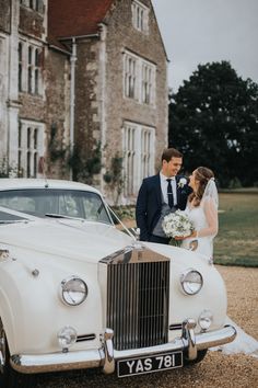 a bride and groom standing in front of an old white wedding car outside a large building