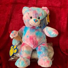 a blue and pink teddy bear sitting on top of a red cloth covered bed sheet