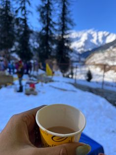 a hand holding a cup of coffee in front of some snow covered mountains and trees