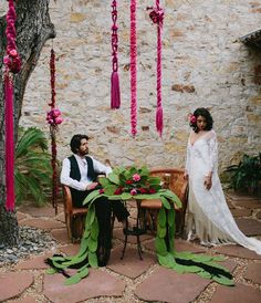 a man and woman sitting at a table in front of a stone wall with flowers hanging from it