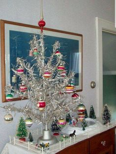 a silver christmas tree with ornaments on it in front of a mirror and framed photograph