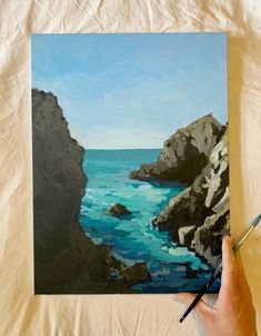 a person holding a paintbrush in front of a painting of the ocean and rocks