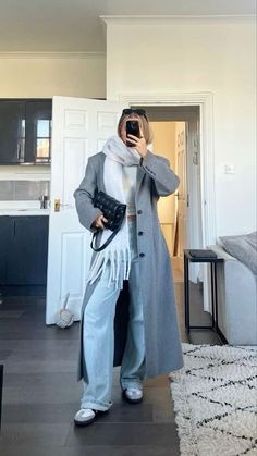 a woman taking a selfie in her living room wearing blue pants and a gray coat