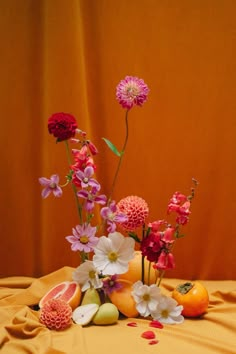 a vase filled with flowers and fruit on top of a yellow table cloth next to an orange wall