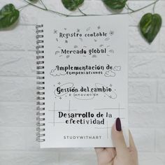 someone holding up a spiral notebook with spanish writing on it and some green plants in the background