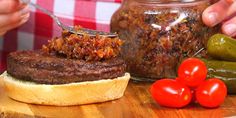 a person is spooning some food out of a jar on a cutting board next to tomatoes and pickles