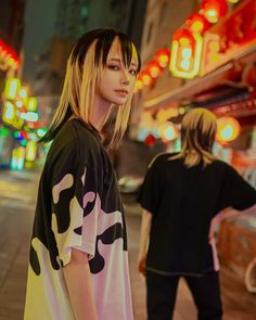 two people standing on the side walk in front of neon signs and buildings at night