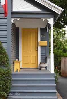 a yellow door sits on the front porch of a gray house with white trim and columns