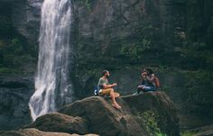 two people sitting on rocks in front of a waterfall