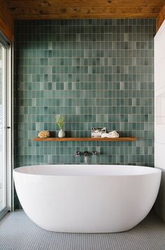 a large white bath tub sitting next to a wooden shelf in a bathroom with green tiles on the walls