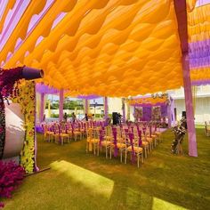 an outdoor wedding setup with purple and yellow decorations on the ceiling, chairs and tables