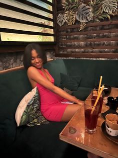 a woman in a pink dress sitting on a green couch next to a table with drinks