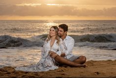 a man and woman sitting on the beach at sunset with waves coming in behind them