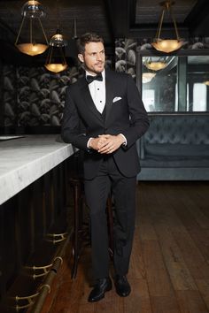 a man in a tuxedo standing next to a bar with lights hanging from the ceiling