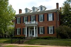 an old brick house with black shutters and white trim on the front porch, surrounded by trees