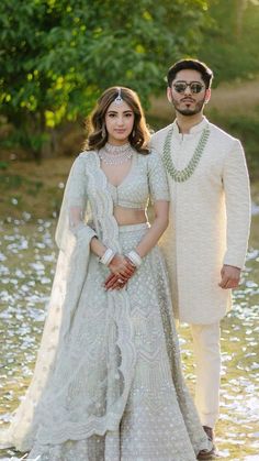 two people standing next to each other in front of some water and trees, wearing white outfits