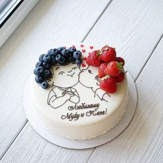 a white cake topped with berries and blueberries on top of a wooden table next to a window