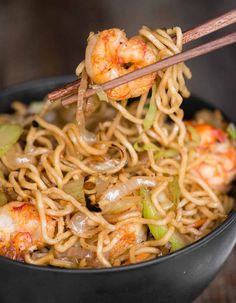 chopsticks holding noodles with shrimp and vegetables in a black bowl on a wooden table