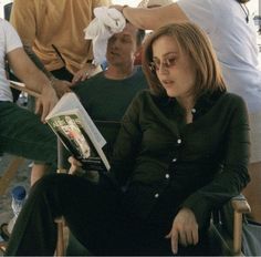 a woman sitting in a chair reading a book with other people standing around behind her