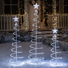 three lighted christmas trees in front of a house with snow and lights on the ground