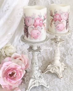 two white candles with pink flowers on them next to a lace tablecloth and curtain