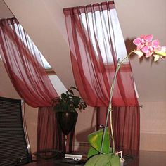 a flower in a vase sitting on a desk next to a window with red drapes