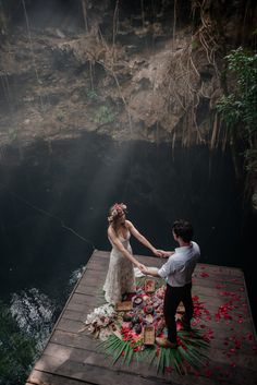 a man and woman holding hands on a dock with flowers in front of the water