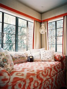 an orange and white day bed in front of two large windows with curtains on both sides