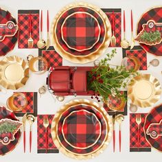 a table set with red and black plaid place mats, gold plates, silverware and greenery