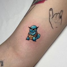 a small tattoo on the arm of a person with a hand in the background and a blue turtle sitting next to it