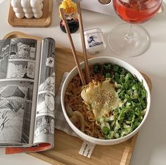 a bowl of noodles and vegetables with chopsticks on a tray next to an open book