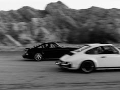 black and white photograph of two cars driving down the road in front of some mountains