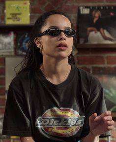 a woman wearing sunglasses standing in front of a brick wall with posters on the wall behind her