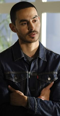 a man with his arms crossed standing in front of a window wearing a denim jacket