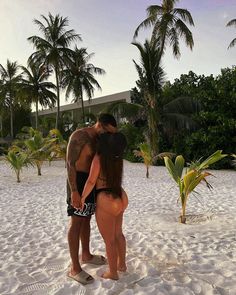 two people standing in the sand with palm trees behind them and one person wearing a black swimsuit