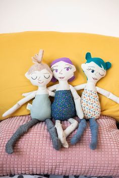 three dolls sitting on top of a bed next to a yellow pillow with polka dots