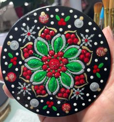 a hand holding a black and red painted round object with green leaves, berries, and dots on it