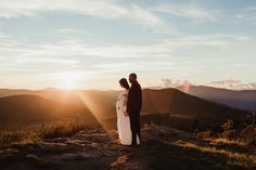 a man and woman standing on top of a mountain looking at the sun in the distance