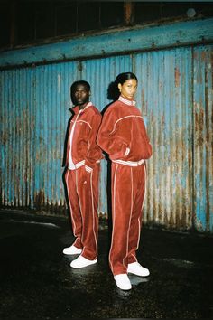 two people in matching red tracksuits standing next to a blue metal wall with peeling paint