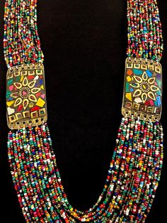 This is a wonderful colorful vintage Naga necklace with two mosaic inlay brass medallions that also serve as connectors for the multi bead strands.  There are 21 strands connected to the top of the mosaic medallion, and 24 rows connected to the bottom of the medallions.   The medallions have a decorative brass sun at center, with their rays having scalloped edges, and a tiny beaded effect frames the center brass circle. The inner circumference of the necklace measures approximately 31". Fastens with a blue disc bead and loop.  Medallions measure approximately 2-1/4" X 1-3/8" Excellent vintage condition.  No issues. Includes gift box and storage pouch. Box 1 Colorful Adjustable Traditional Necklaces, Traditional Multicolor Polished Beads Jewelry, Spiritual Multicolor Beaded Necklaces For Rituals, Traditional Colorful Necklaces For Festival, Traditional Multicolor Long Beaded Necklace, Traditional Multicolor Jewelry With Gold Beads, Traditional Multicolor Beads For Jewelry Making, Multicolor Traditional Beads For Jewelry Making, Traditional Beaded Necklaces With Colorful Beads For Festival